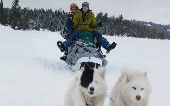 a person sits on a sled and smiles while another stands at the back of the sled, all being pulled by a team of dogs, two of which you can see in the foreground